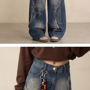 May include: Blue denim wide-leg jeans with distressed detailing and embroidered sun, star, and heart designs. Features a brown leather charm and silver keychain. The jeans have a high waist and a relaxed fit.