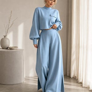 Puede incluir: Un conjunto de dos piezas azul claro que consta de una parte superior corta de manga larga con cuello alto, un peque&ntilde;o bolsillo en el pecho y mangas voluminosas, combinado con pantalones anchos de tiro alto con costuras y aberturas laterales.