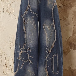 May include: Blue denim jeans with a wide leg design. The jeans feature embroidered celestial designs, including stars, a sun, and a moon. The edges of the jeans have a distressed, frayed detail.