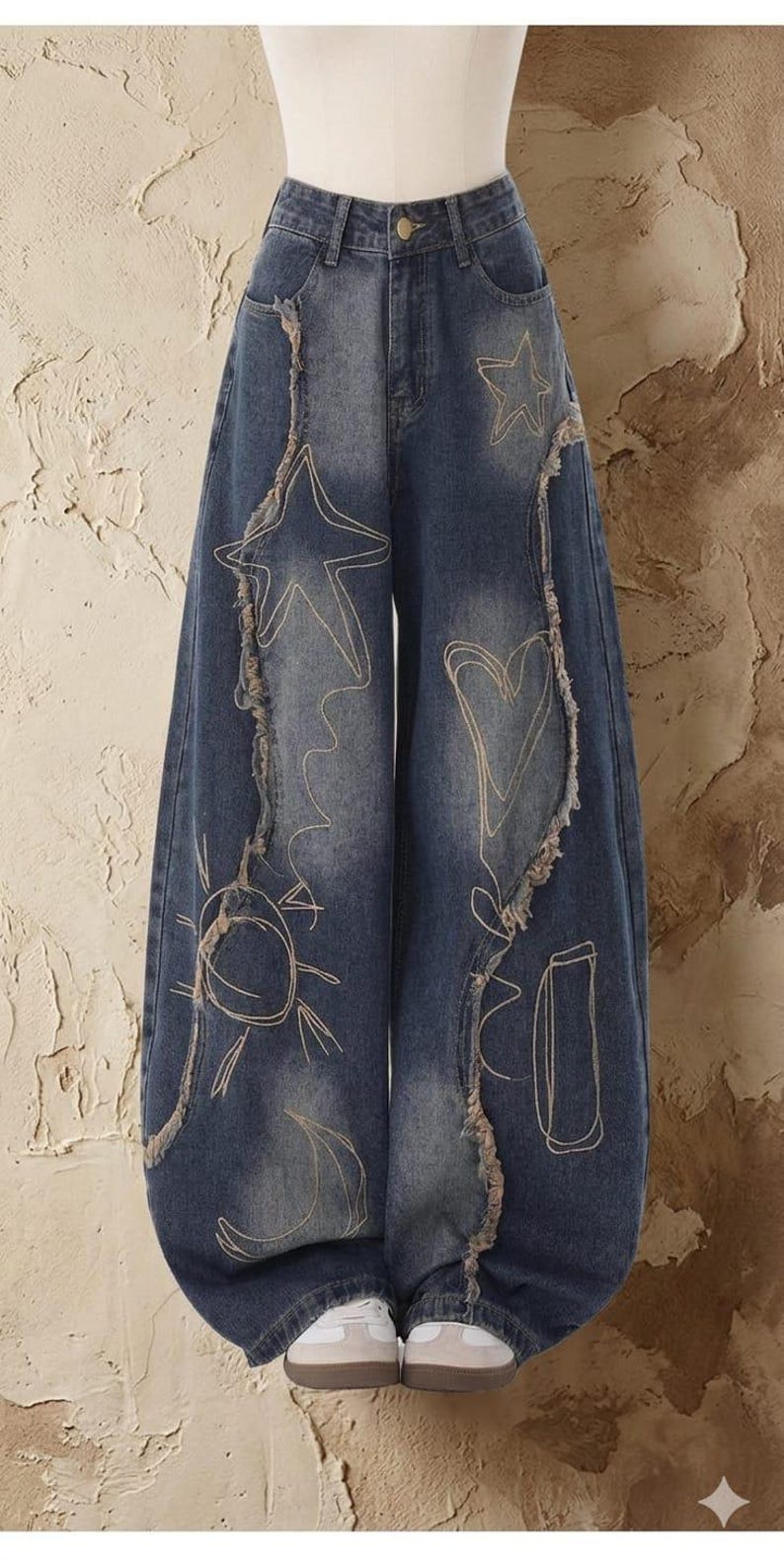 May include: Blue denim jeans with a wide leg design. The jeans feature embroidered celestial designs, including stars, a sun, and a moon. The edges of the jeans have a distressed, frayed detail.