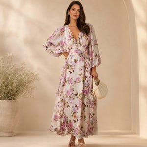 May include: A long-sleeved maxi dress featuring a white base with a floral pattern in shades of purple, pink, and green. The dress has a deep V-neckline, a cinched waist, and vertical ruffled detailing along the skirt, paired with a small beige handbag.