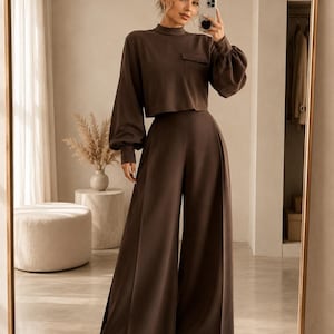 Puede incluir: Un conjunto de dos piezas en color marr&oacute;n oscuro que consta de una parte superior corta de manga larga con cuello alto, pu&ntilde;os fruncidos y un bolsillo en el pecho, junto con pantalones de talle alto y pernera ancha de silueta estructurada.