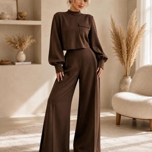 Puede incluir: Un conjunto de dos piezas de color marr&oacute;n oscuro que consta de un top corto de manga larga con cuello alto y un bolsillo en el pecho, combinado con pantalones de pierna ancha y cintura alta con una raya marcada en cada pierna y zapatos blancos.