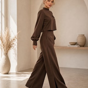 Puede incluir: Un conjunto de dos piezas de color marr&oacute;n oscuro que consta de un top corto de manga larga con cuello alto y un peque&ntilde;o bolsillo en el pecho, combinado con pantalones de tiro alto y pernera ancha con aberturas laterales y zapatos blancos.