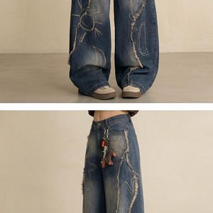 May include: Wide-leg, distressed denim jeans in a medium wash. The jeans feature decorative stitching and graphic designs of stars, suns, and hearts. Frayed edges and a keychain add detail to the casual, yet stylish, design.