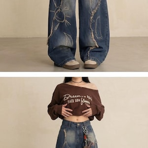 May include: Wide-leg, distressed denim jeans with embroidered star, sun, and heart designs. The jeans have a faded wash and frayed edges. A brown off-the-shoulder sweatshirt with the text "Dream of life, Still life flower" is also visible.
