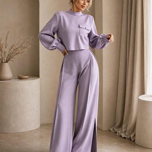 Puede incluir: Un conjunto de dos piezas en color lavanda que consta de una blusa de manga larga con cuello alto y un bolsillo en el pecho, junto con pantalones de talle alto y pernera ancha con pliegues verticales y aberturas laterales, con zapatos blancos.