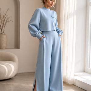Puede incluir: Un conjunto de dos piezas en color azul claro que consta de una parte superior corta de manga larga con cuello alto y un bolsillo en el pecho, junto con pantalones de talle alto y pernera ancha con aberturas laterales y bolsillos.