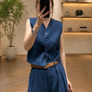 May include: A dark blue sleeveless vest with a V-neckline and five matching buttons down the front, paired with matching high-waisted pleated trousers and a tan leather belt featuring a gold-toned buckle at the center of the waistline.