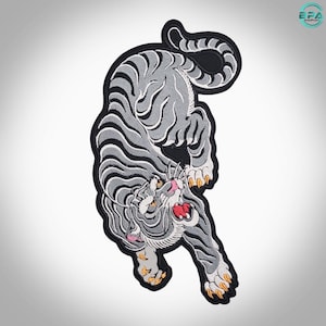 May include: An embroidered patch featuring a gray tiger with black stripes in a downward pouncing position. The tiger has a white face, a pink nose, an open mouth with red interior, and yellow claws. The entire design is outlined in black thread.