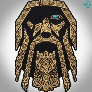 Può includere: Una toppa ricamata nera con un design color oro di ispirazione norrena che raffigura una figura barbuta con intricati motivi a intreccio. Il disegno include due uccelli sopra la barba e un dettaglio di un occhio blu brillante in alto.