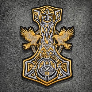 May include: An embroidered patch featuring a silver and gold Thor's hammer design. The hammer is adorned with intricate knotwork, a central triquetra symbol, and two golden ravens with outstretched wings flanking the upper section of the handle.