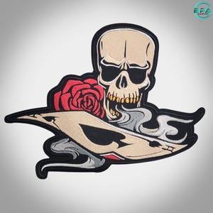 May include: An embroidered patch featuring a beige skull with black eye sockets and yellow teeth, positioned above a red rose and a stylised playing card with a black spade symbol, all surrounded by swirling grey smoke on a black border.