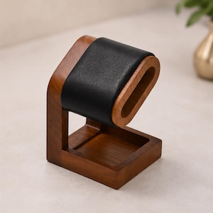May include: A watch display stand featuring a brown wooden base and a vertical support arm. The top section consists of a curved, oval-shaped wooden frame wrapped in a smooth, black leather-like material designed to hold a wristwatch securely.
