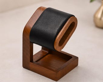 May include: A watch display stand featuring a brown wooden base and a vertical support arm. The top section consists of a curved, oval-shaped wooden frame wrapped in a smooth, black leather-like material designed to hold a wristwatch securely.
