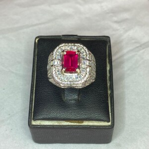 May include: An elaborate ring with a central, rectangular, deep red gemstone. The gemstone is encircled by smaller, clear stones, set in a silver-coloured metal band. The ring is presented on a black jewellery box.
