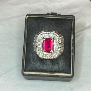 May include: A silver ring featuring a large, rectangular, bright red gemstone at its centre, encircled by smaller, clear stones. The ring is presented in a square black box with a white interior.