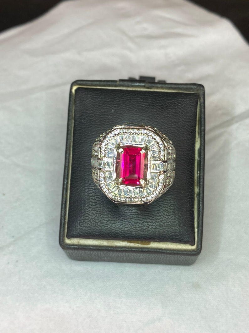 May include: A silver ring featuring a large, rectangular, bright red gemstone at its centre, encircled by smaller, clear stones. The ring is presented in a square black box with a white interior.