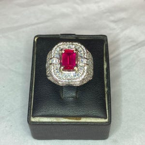 May include: An elaborate ring featuring a vibrant, rectangular red gemstone. The central stone is framed by a halo of smaller, clear gems. The ring's band is also adorned with clear stones, creating a dazzling appearance. The ring is presented on a black display box.