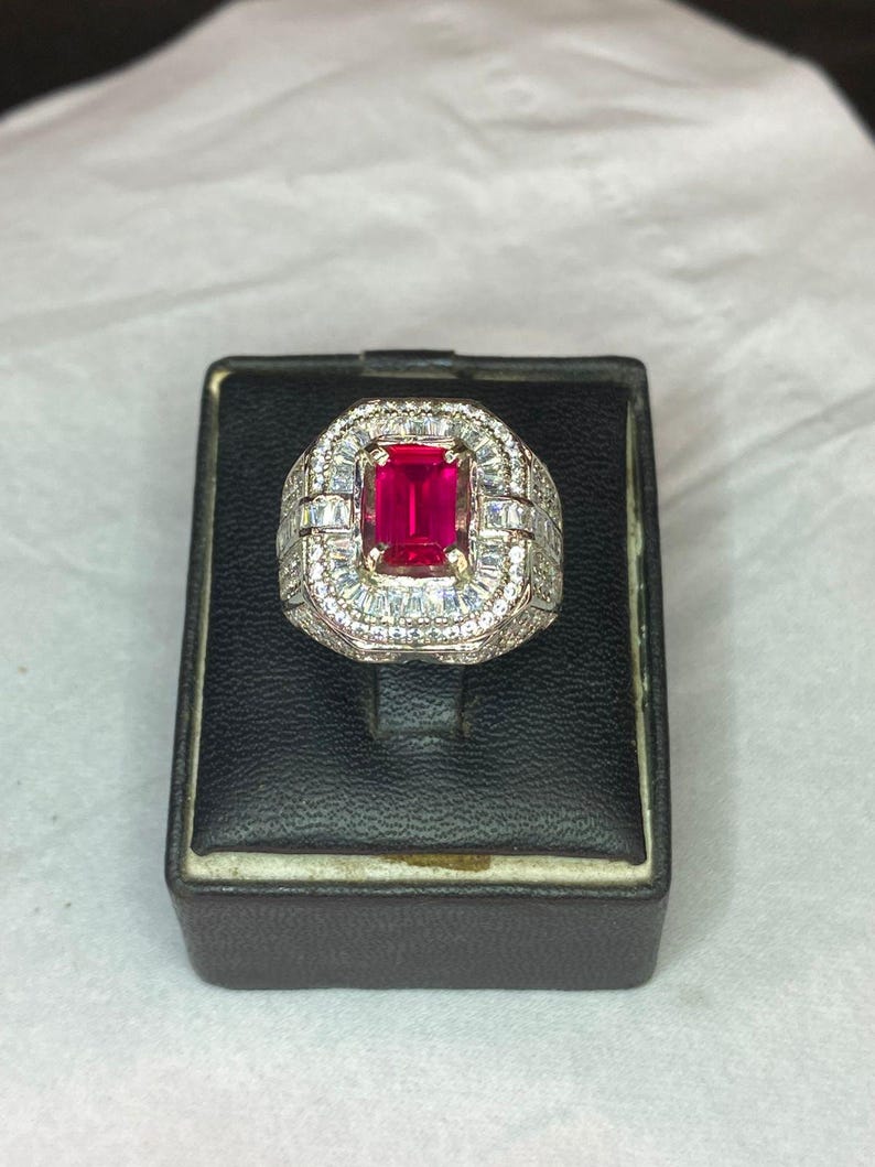May include: An elaborate ring featuring a vibrant, rectangular red gemstone. The central stone is framed by a halo of smaller, clear gems. The ring's band is also adorned with clear stones, creating a dazzling appearance. The ring is presented on a black display box.