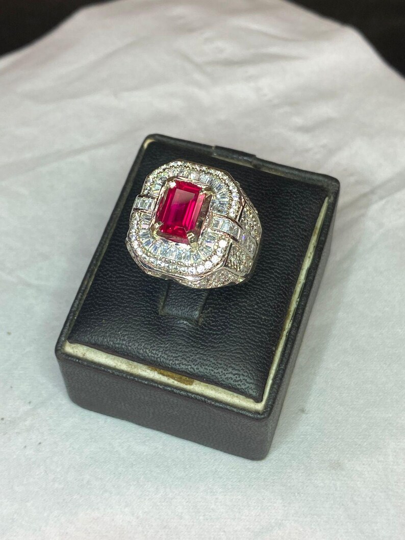 May include: A silver ring featuring a large, rectangular, red gemstone. The ring is encircled by smaller, clear gemstones. The ring is presented on a black jewellery box.