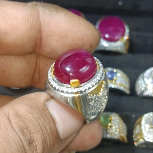 May include: A silver ring with a large, oval, deep red gemstone. The gemstone is surrounded by a halo of small, clear stones. The ring has gold accents and is held by a hand. Other rings are visible in the background.