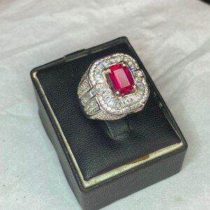 May include: A silver ring featuring a large, rectangular, red gemstone. The central stone is encircled by smaller, clear stones. The ring is presented on a black display box.