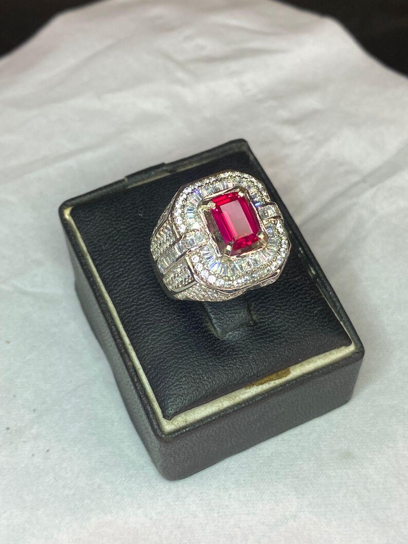 May include: A silver ring featuring a large, rectangular, red gemstone. The central stone is encircled by smaller, clear stones. The ring is presented on a black display box.