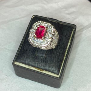 May include: A silver ring featuring a large, rectangular, red gemstone. The ring is encircled by smaller, clear gemstones. The ring is presented on a black jewellery box.