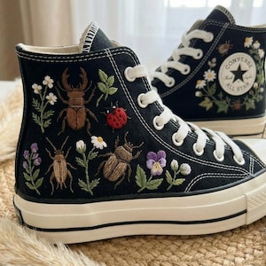 Könnte beinhalten: Schwarze High-Top-Sneaker mit gestickten Insekten und floralen Designs. Die Sneaker haben weiße Schnürsenkel, eine cremefarbene Sohle und das Converse All Star-Logo. Die Stickerei umfasst Käfer, einen Marienkäfer und verschiedene Blumen.