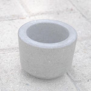 May include: A small, light gray, cylindrical mortar with a wide opening. The mortar has a rough, textured surface and is set against a light-colored, textured background. The mortar is likely made of stone or concrete.
