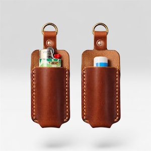 Pode incluir: Duas capas de isqueiro em couro castanho com ferragens em latão. Uma capa contém um isqueiro BIC verde e branco, a outra um protetor labial branco e azul. Ambas as capas têm um design costurado e um anel de latão para fixação.