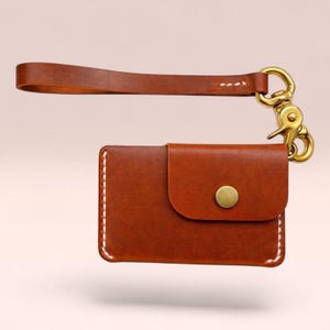 Pode incluir: Um porta-cartões de couro castanho com uma alça de pulso e um fecho dourado. O porta-cartões tem uma aba com um fecho de botão dourado e costuras brancas nas bordas. A alça de pulso é presa ao porta-cartões com um fecho dourado.
