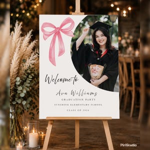 Puede incluir: Un letrero de bienvenida para una fiesta de graduación sobre un caballete de madera. El letrero presenta un lazo rosa de acuarela y una foto de una graduada con birrete y toga. El texto dice "Bienvenida a Ava Williams, Fiesta de Graduación, Sunshine Elementary School, Clase de 2026."