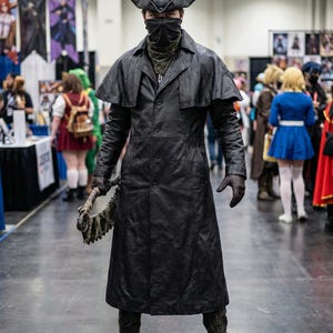 May include: A person in a black leather coat and hat, with a face covering, holding a weapon. The costume is reminiscent of a character from a video game or movie. The setting appears to be a convention or event.