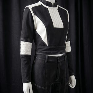 Puede incluir: Un conjunto de dos piezas en blanco y negro sobre un maniqu&iacute;. La parte superior tiene un cuello alto y detalles en blanco. Los pantalones son negros con costuras blancas. El conjunto tiene un dise&ntilde;o futurista.