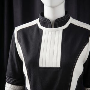 Puede incluir: Una prenda tipo t&uacute;nica en blanco y negro con cuello alto y mangas largas. El dise&ntilde;o presenta paneles y detalles blancos, incluyendo un panel vertical en el pecho y detalles acolchados en hombros y mangas. La prenda se exhibe en un maniqu&iacute;.