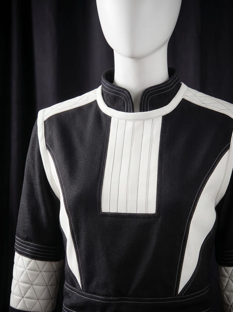 Puede incluir: Una prenda tipo t&uacute;nica en blanco y negro con cuello alto y mangas largas. El dise&ntilde;o presenta paneles y detalles blancos, incluyendo un panel vertical en el pecho y detalles acolchados en hombros y mangas. La prenda se exhibe en un maniqu&iacute;.