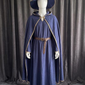 Puede incluir: Una capa con capucha de color azul oscuro con ribete dorado, un sombrero puntiagudo a juego con ribete dorado y un vestido largo de manga larga. Un cinturón de cuero marrón con hebilla metálica se ajusta a la cintura del vestido sobre un maniquí.