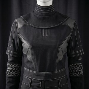May include: Black jumpsuit with white stitching details. Features a high, ribbed collar, leather-like panels, and geometric patterns on the sleeves. The jumpsuit has a belt with a buckle.
