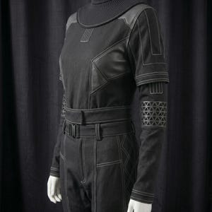 May include: Black jumpsuit with a high, ribbed collar and long sleeves. The jumpsuit features leather-like accents on the shoulders and upper arms, with geometric patterns on the lower sleeves. White stitching details the seams and pockets.