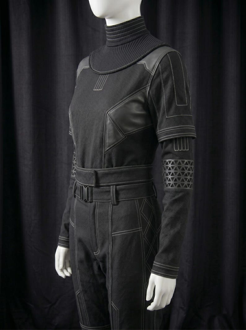 May include: Black jumpsuit with a high, ribbed collar and long sleeves. The jumpsuit features leather-like accents on the shoulders and upper arms, with geometric patterns on the lower sleeves. White stitching details the seams and pockets.