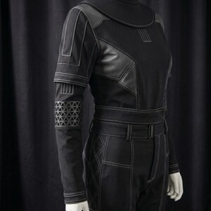 May include: Black jumpsuit with white stitching details. The outfit features a high collar, leather accents, and geometric patterns on the sleeves. The pants have a belt and a tailored fit, creating a futuristic or cosplay aesthetic.