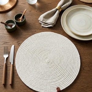 May include: A white, circular woven placemat with a brown leather accent is the focal point of this table setting. The setting includes a fork, knife, plates, napkin, and small bowls with wooden spoons, all arranged on a wooden table.