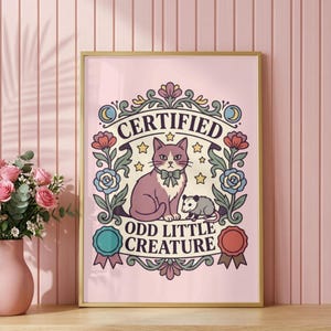 May include: A rectangular wall art print in a gold frame featuring a central illustration of a purple cat with a bow tie and a small opossum. The text reads CERTIFIED ODD LITTLE CREATURE, surrounded by floral motifs, stars, and two circular ribbons.