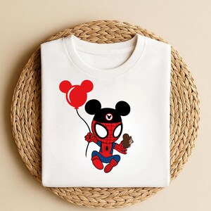 May include: White t-shirt featuring a cartoon character combining elements of Spider-Man and Mickey Mouse. The character holds a red balloon and a chocolate treat. The design is in red, blue, black, and white.