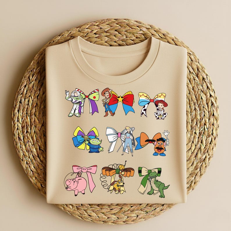 Puede incluir: Una camiseta beige con lazos de personajes de dibujos animados coloridos. Los lazos incluyen a Buzz Lightyear, Woody y otros personajes. La camiseta se muestra sobre una superficie tejida circular.