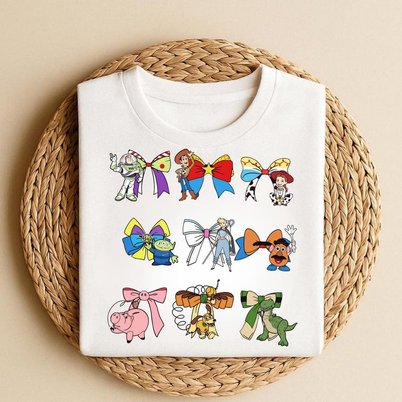Puede incluir: Camiseta blanca con dise&ntilde;os de personajes de dibujos animados con lazos. Los dise&ntilde;os incluyen a Buzz Lightyear, Woody, Jessie y otros personajes. La camiseta se muestra sobre una superficie circular tejida.
