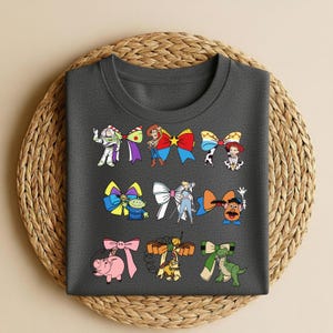 Puede incluir: Una camiseta gris oscuro con lazos de personajes de dibujos animados coloridos. Los lazos incluyen personajes como Buzz Lightyear y Woody. La camiseta se muestra sobre una estera circular tejida.