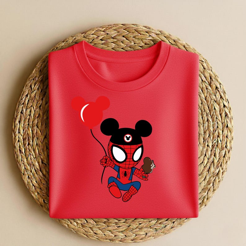 May include: A folded red t-shirt featuring a cartoon character that is a hybrid of Spider-Man and Mickey Mouse. The character is holding a red balloon and a chocolate ice cream cone. The shirt is on a woven, circular placemat.